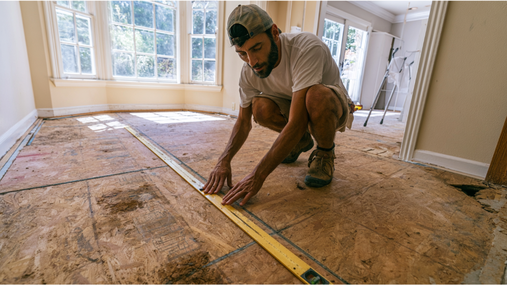 Contractor checking exposed plywood subfloor after tile removal using a straightedge and level before installing new flooring.