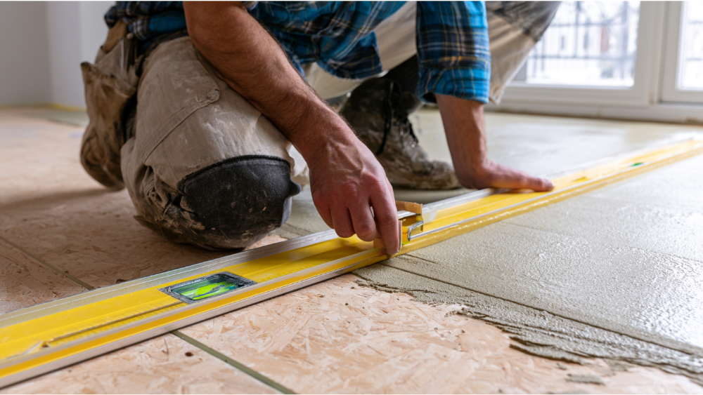 Contractor inspecting and leveling a plywood subfloor before tile installation using straightedge, level, and self-leveling compound.