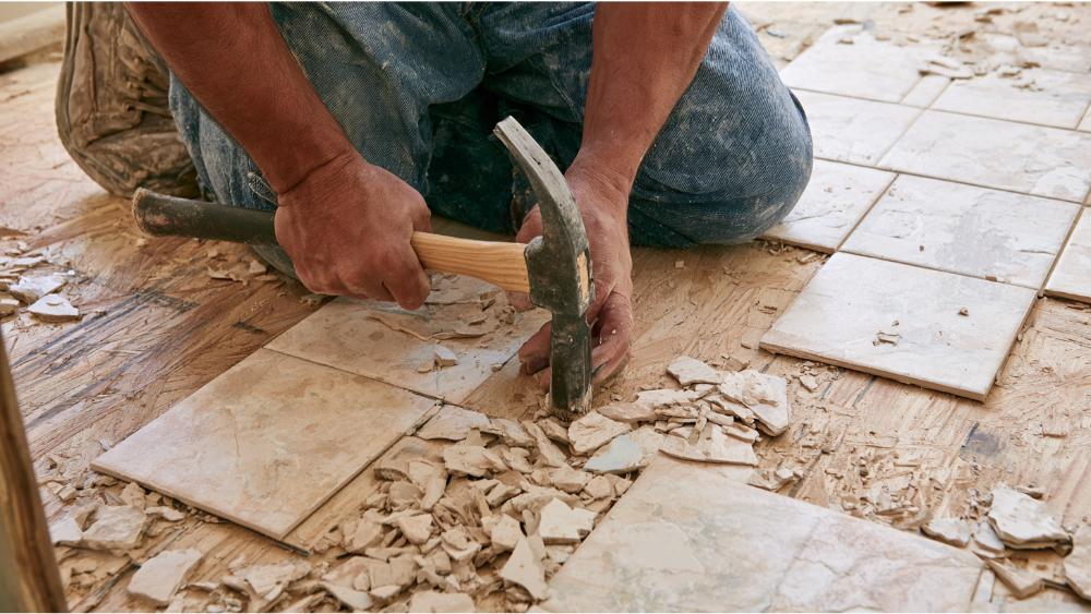 Contractor lifting ceramic tiles carefully with a pry bar at a shallow angle to protect the wood subfloor underneath.